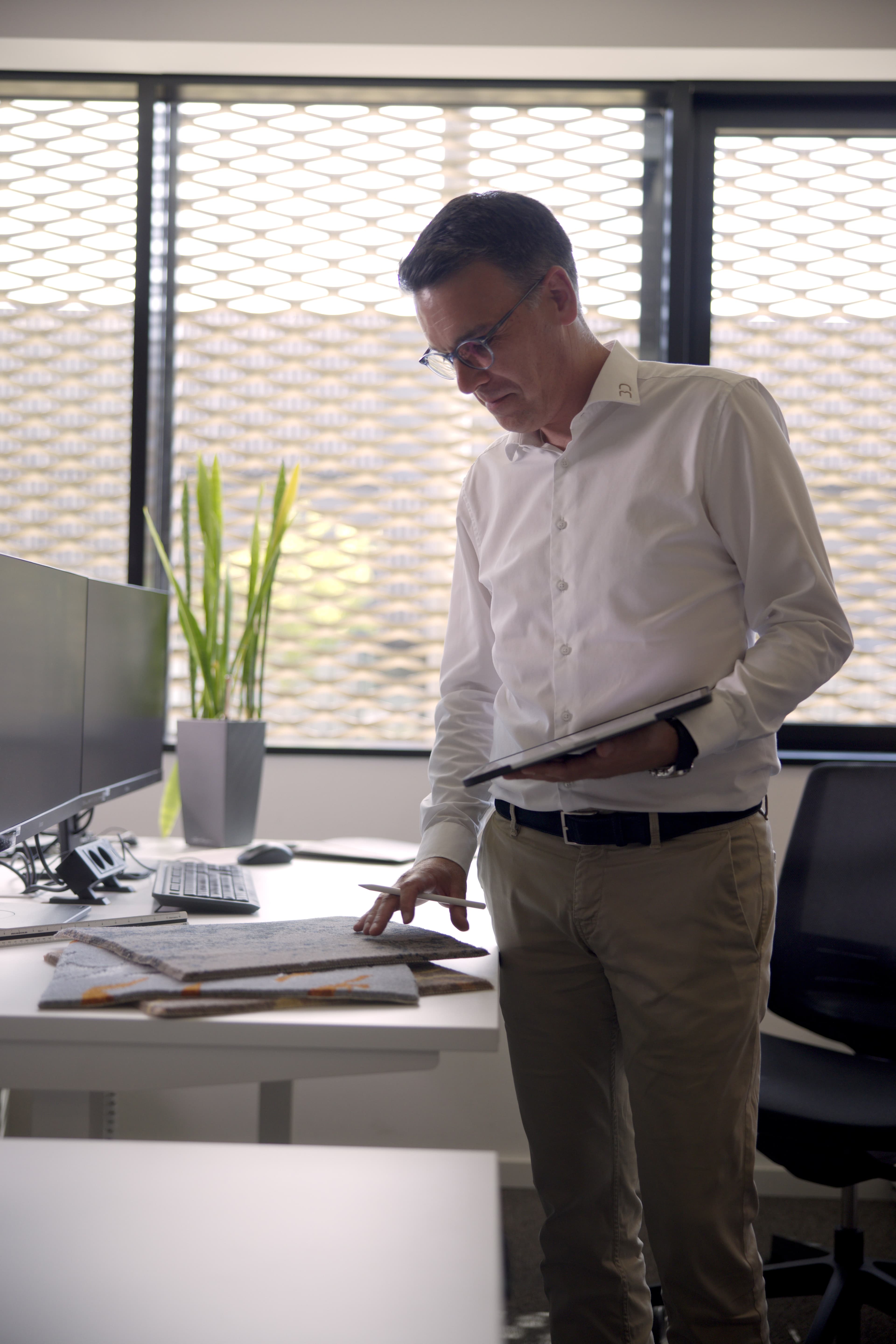 Mann in weißem Hemd betrachtet Baupläne auf einem Schreibtisch in einem hellen Büro und hält dabei ein Tablet in der Hand.